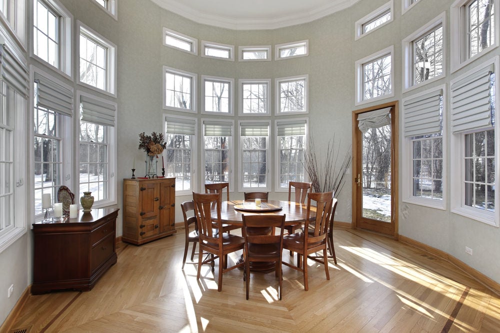 A transitional sunroom with a high ceiling, wooden furniture and dirty white walls.